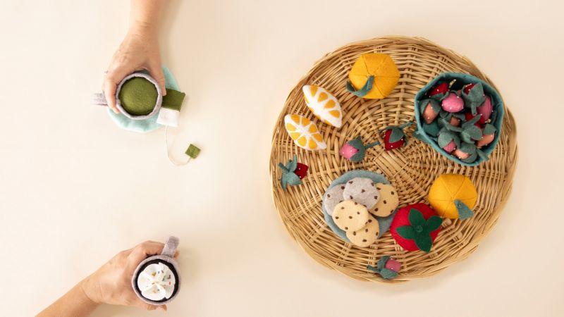 Two hands with felt drinks near a wicker tray of colorful felt fruits, cookies, and sweets on a light surface.