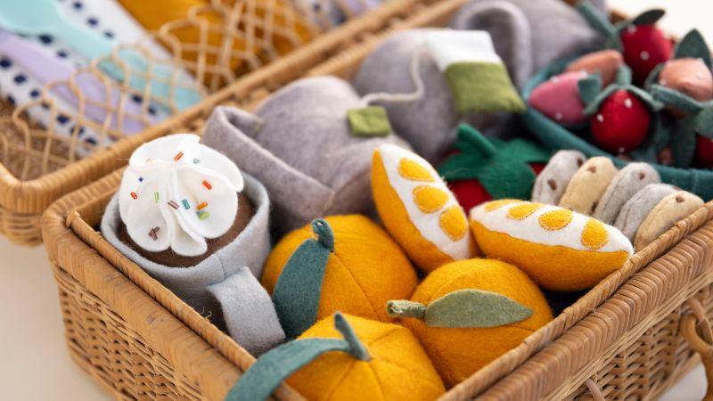 A basket filled with colorful, felt plush fruits, vegetables, and a cup with whipped cream.