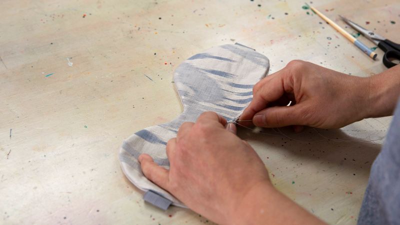 Close-up of hands sewing a gray fabric piece on a table with scissors and paintbrushes in the background.