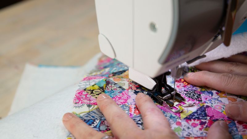 Close-up of hands guiding fabric under a sewing machine needle, stitching a colorful patchwork quilt.