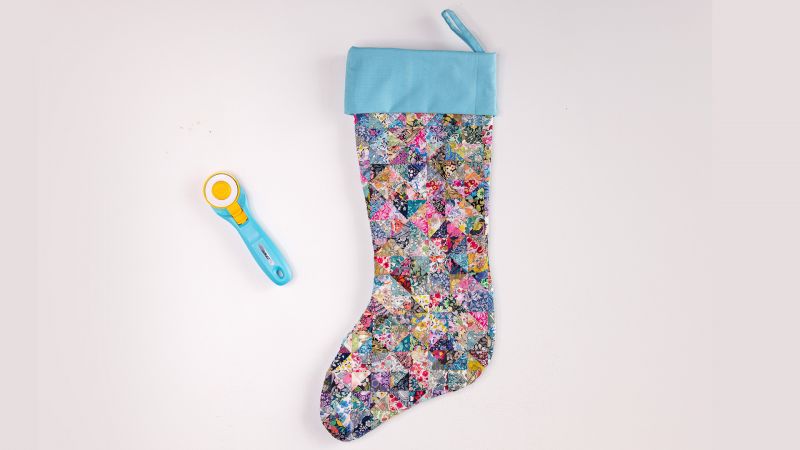 Colorful patchwork Christmas stocking with a light blue cuff beside a blue fabric rotary cutter on a white background.