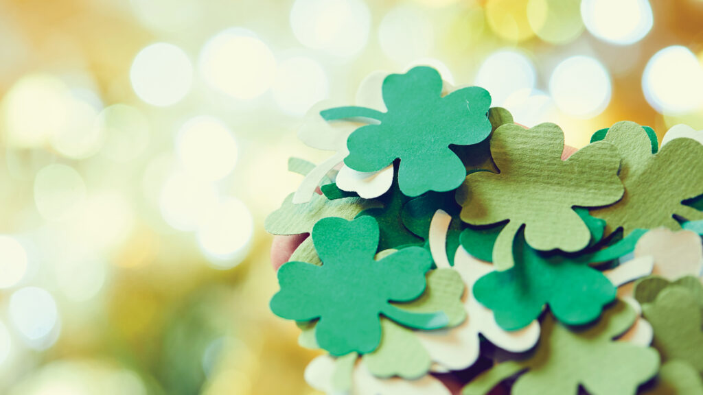 A hand holds a bunch of green paper shamrocks against a blurred, brightly lit background.
