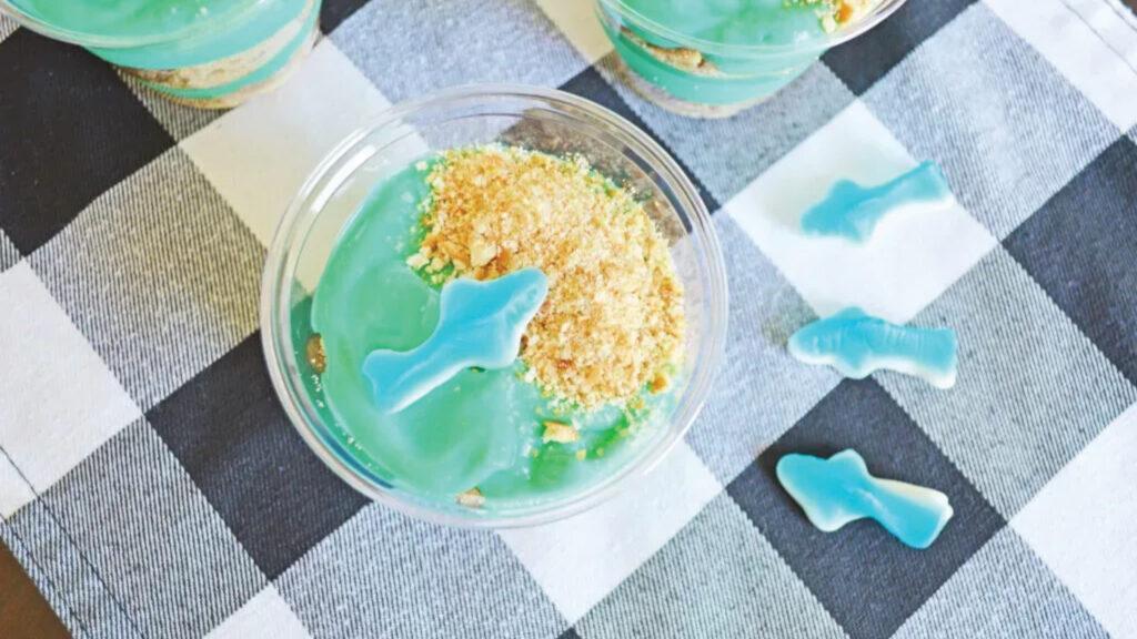 A cup of blue pudding with cookie crumbs and gummy sharks on a checkered tablecloth.