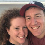 A smiling couple takes a close-up selfie on the beach, with the ocean and sand visible in the background.