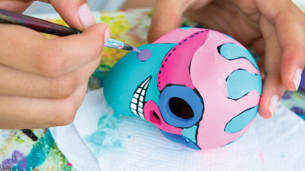 A hand paints a colorful sugar skull with a brush on a white paper towel.
