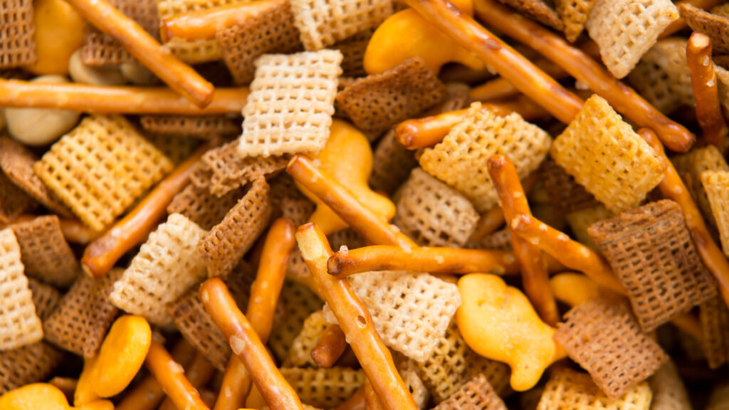 A close-up of a snack mix with pretzel sticks, Chex cereal pieces, and goldfish crackers.