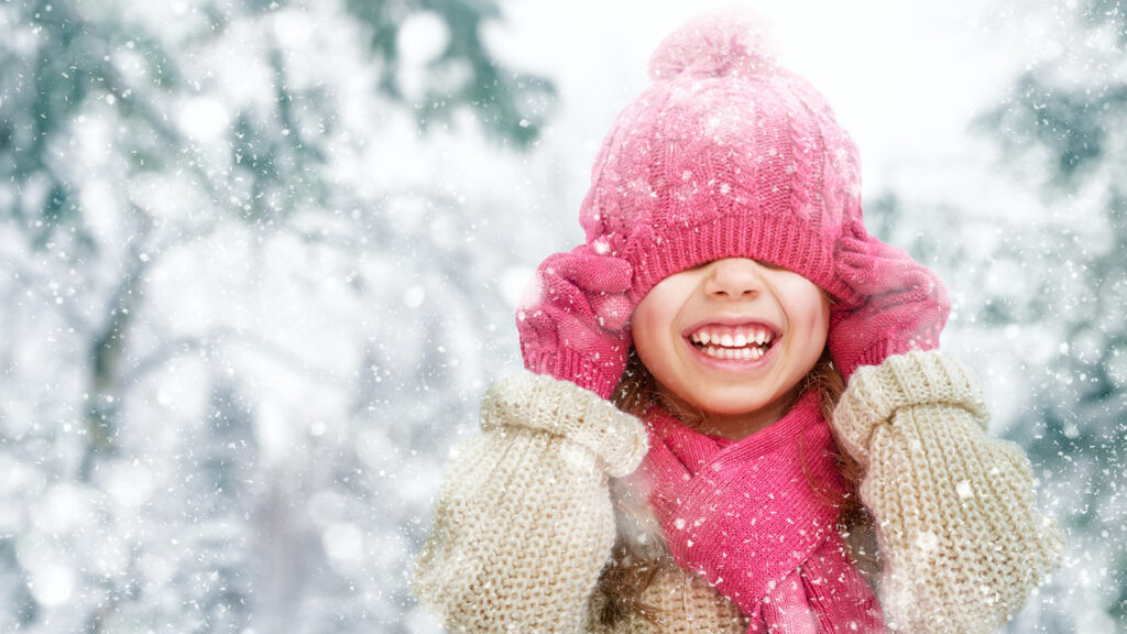 Child smiling in a snowy forest, wearing a pink hat, scarf, and gloves, pulling the hat over their eyes, with snow falling.