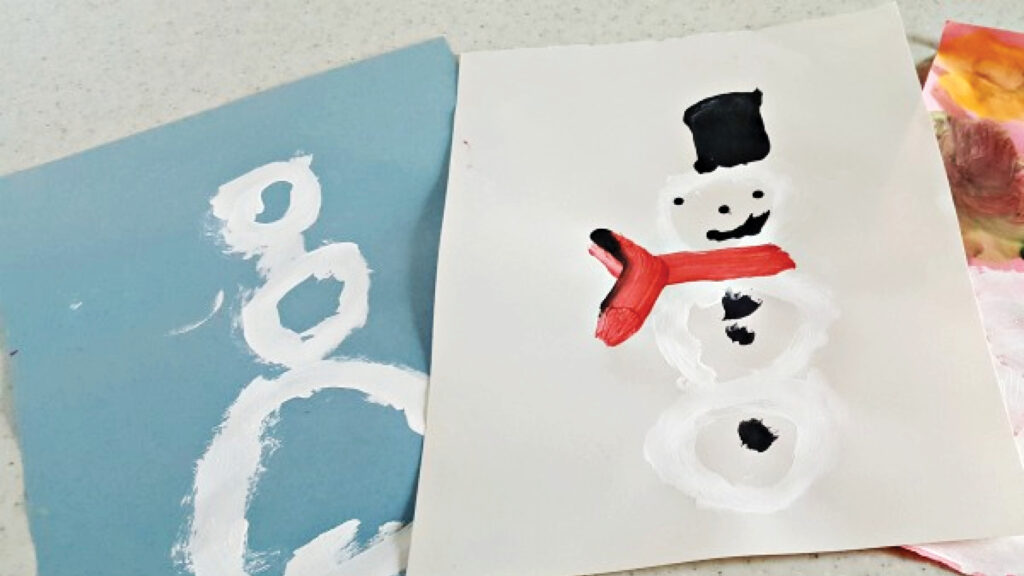 Two child-painted snowmen on paper, one abstract on blue paper, one detailed with hat and scarf on white paper.