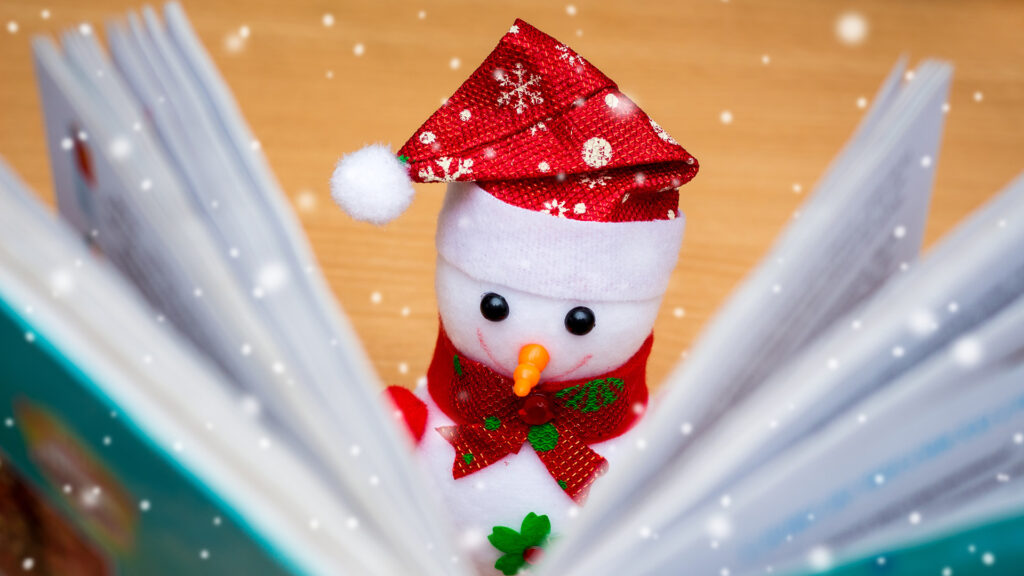 A snowman figurine in a red hat and scarf "reads" an open book, surrounded by falling snow indoors.