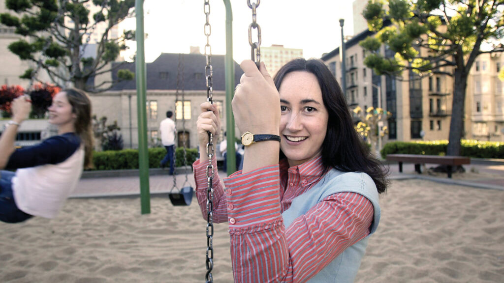 A woman smiles on a swing set in a playground, with another person swinging in the background.