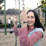 A woman smiles on a swing set in a playground, with another person swinging in the background.
