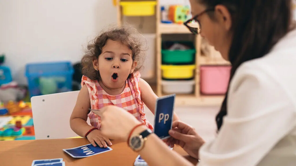 A child and adult practice letters with flashcards at a table in a colorful classroom.