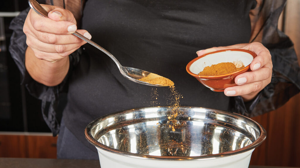 Person pouring ground cinnamon from a spoon into a metal mixing bowl, holding a small bowl of cinnamon.