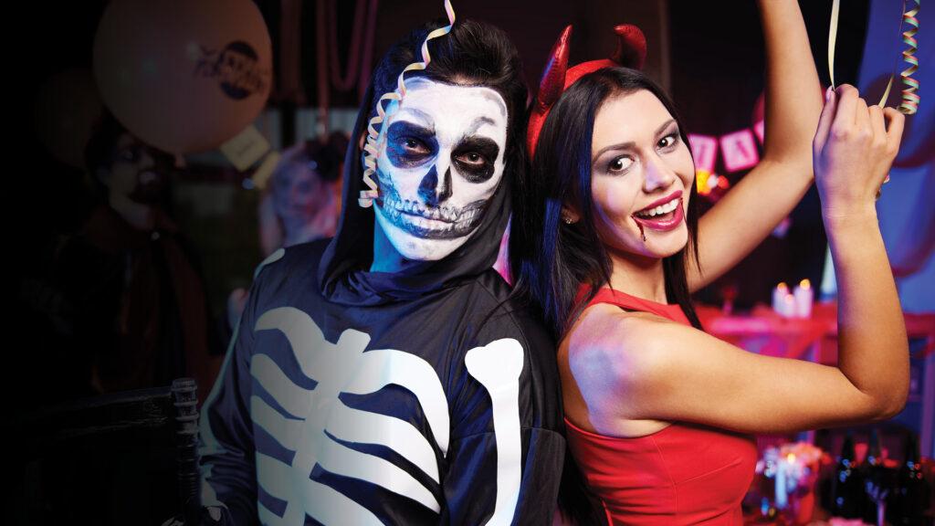 Two people in Halloween costumes: one as a skeleton, the other as a devil, smiling at a festive party.
