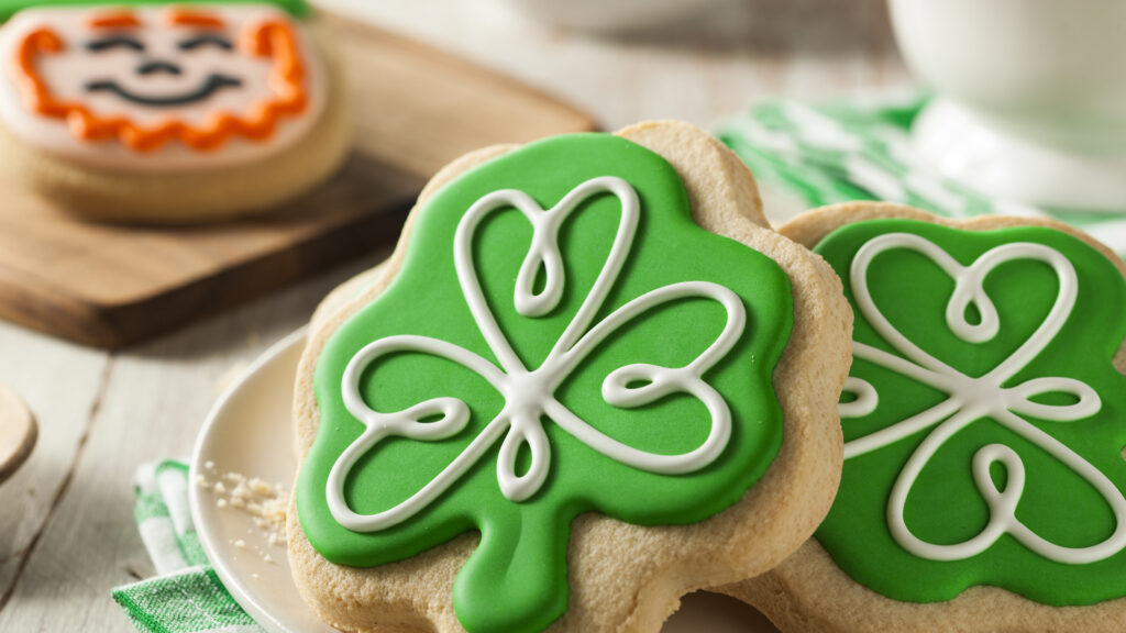 Shamrock-shaped sugar cookies with green and white icing on a plate, with a pumpkin cookie in the background.