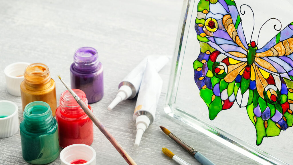 Glass painting of a colorful butterfly with paint jars, brushes, and two paint tubes on a gray surface.