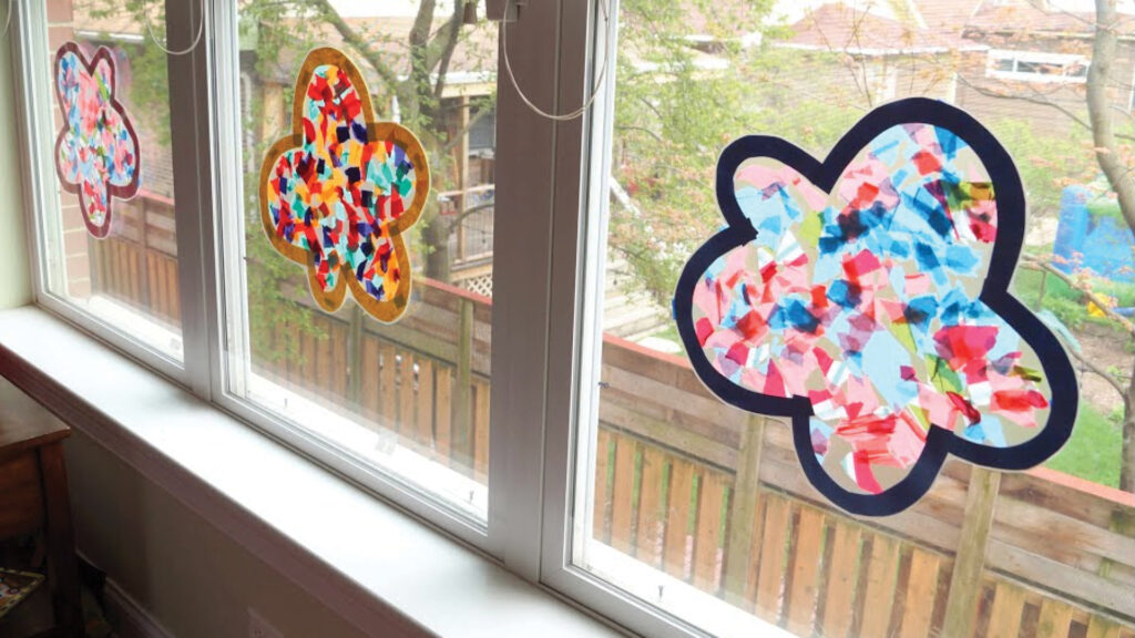 Three colorful, flower-shaped suncatchers are displayed on a window, with a yard visible outside.