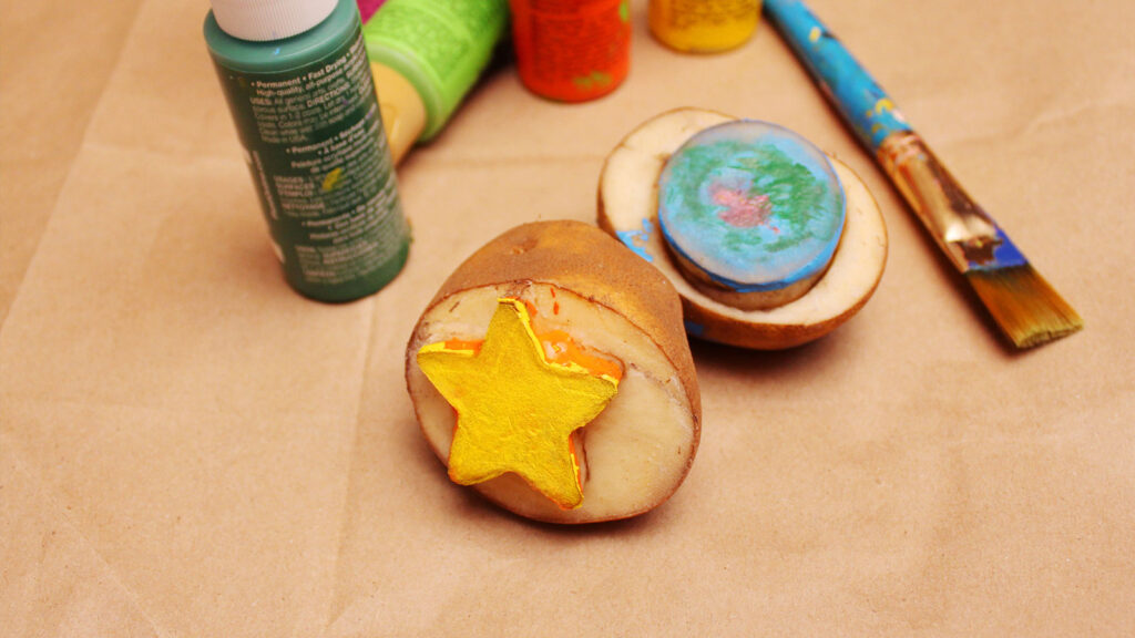 Potato stamps with a yellow star and blue-green circle, paint bottles, and a brush on brown paper.