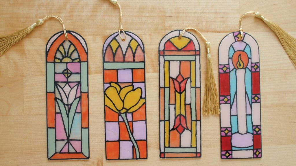 Four colorful stained glass bookmarks with tassels on a wooden surface. Designs feature floral and geometric patterns.