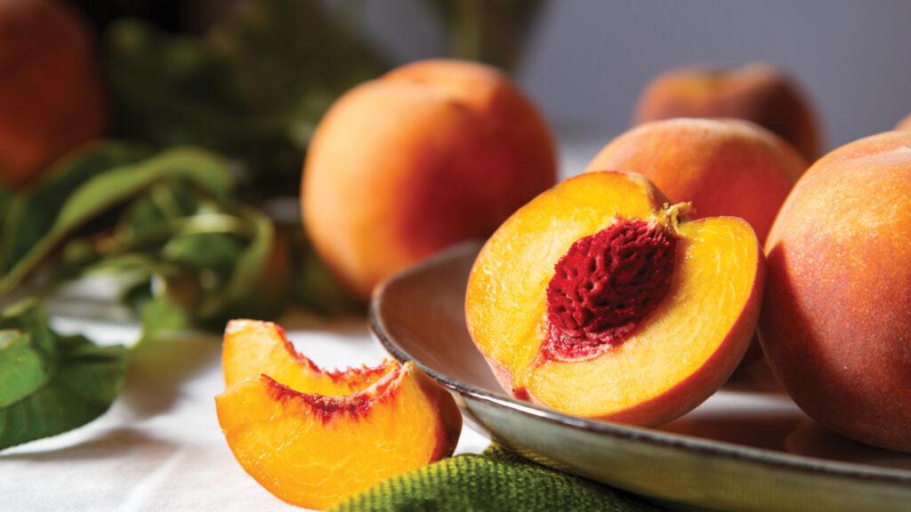 A close-up of fresh peaches, one halved to show the pit, on a plate with green leaves and a peach slice.