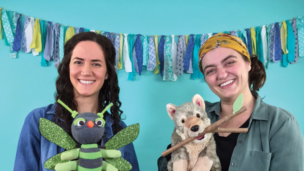 Two women smiling, one holding a dragonfly puppet, the other a wolf puppet, with colorful garland behind them.