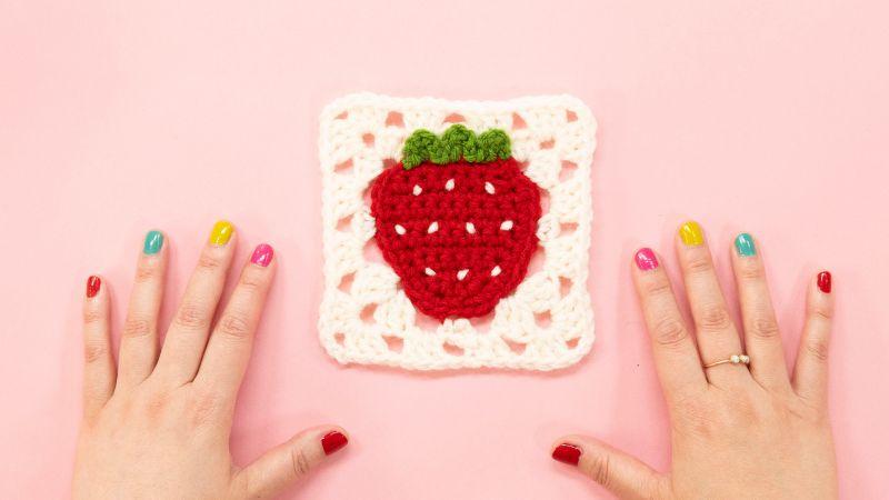 Hands with colorful nails framing a crocheted strawberry square on a pink background.
