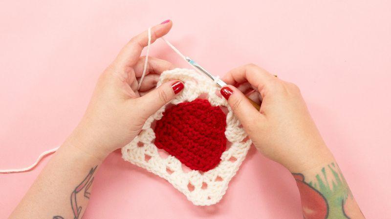 Hands crocheting a white square with a red heart in the center on a pink background.