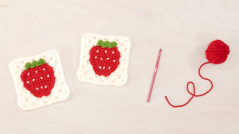 Two crocheted strawberry granny squares, a pink crochet hook, and a ball of red yarn on a light surface.