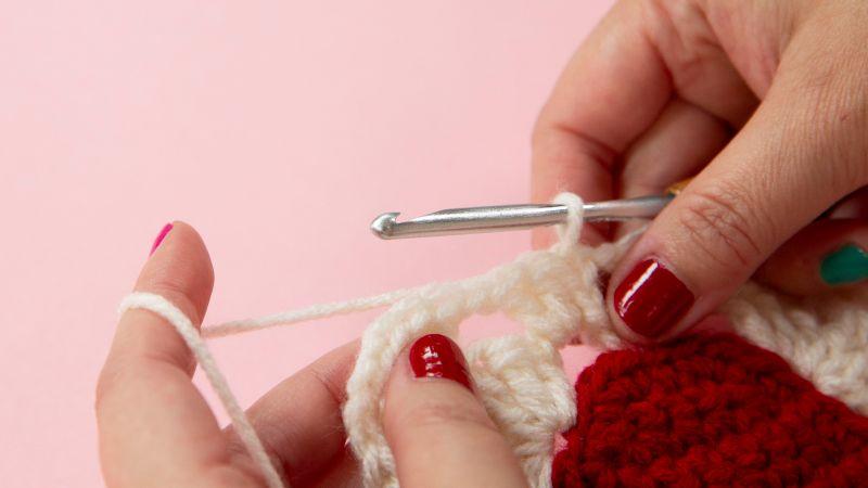 Hands with red nail polish crochet with white yarn and a metal hook against a pink background.