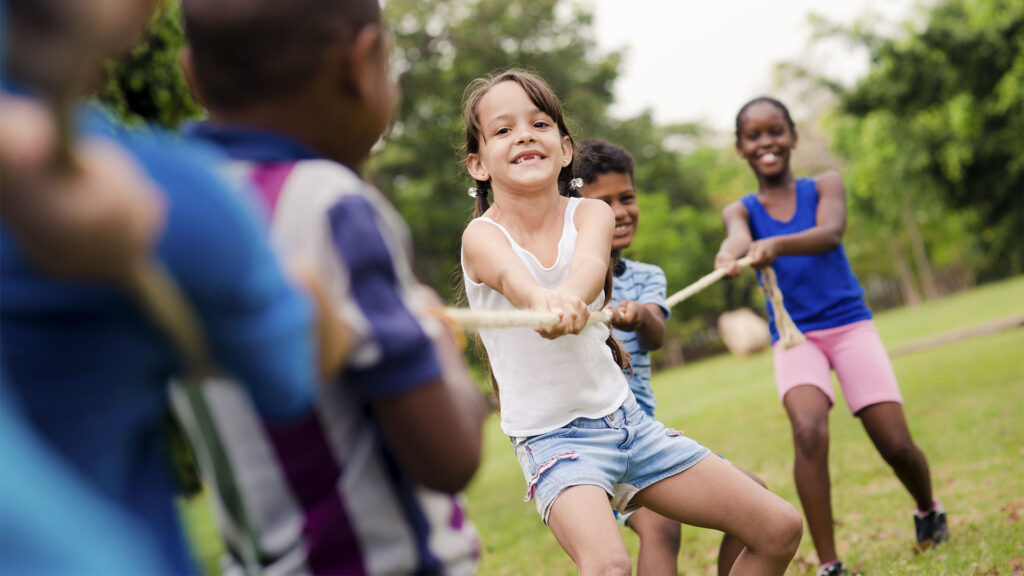 Children playing tug-of-war outside on a grassy field, smiling and pulling the rope energetically on a sunny day.