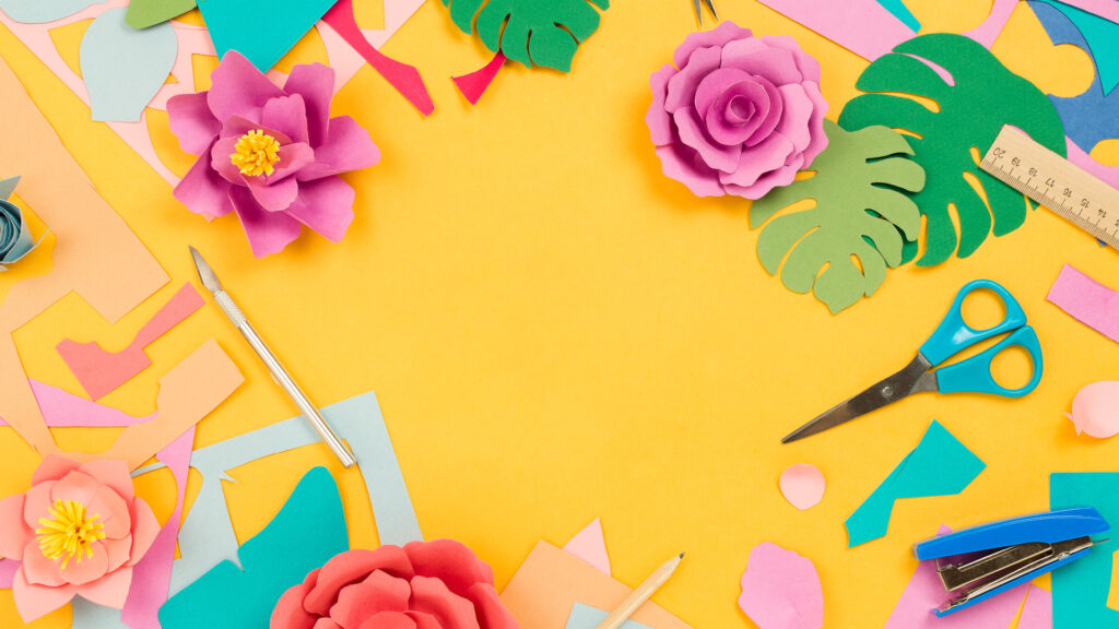 Colorful paper flowers, scissors, ruler, and craft supplies arranged on a yellow background.