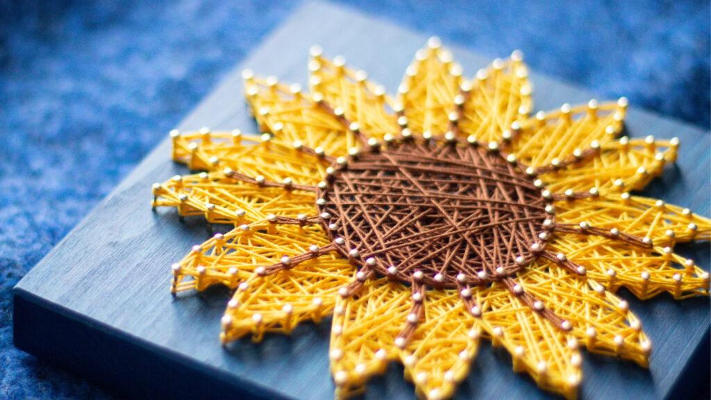 A sunflower made from yellow and brown thread woven on nails placed on a wooden board.