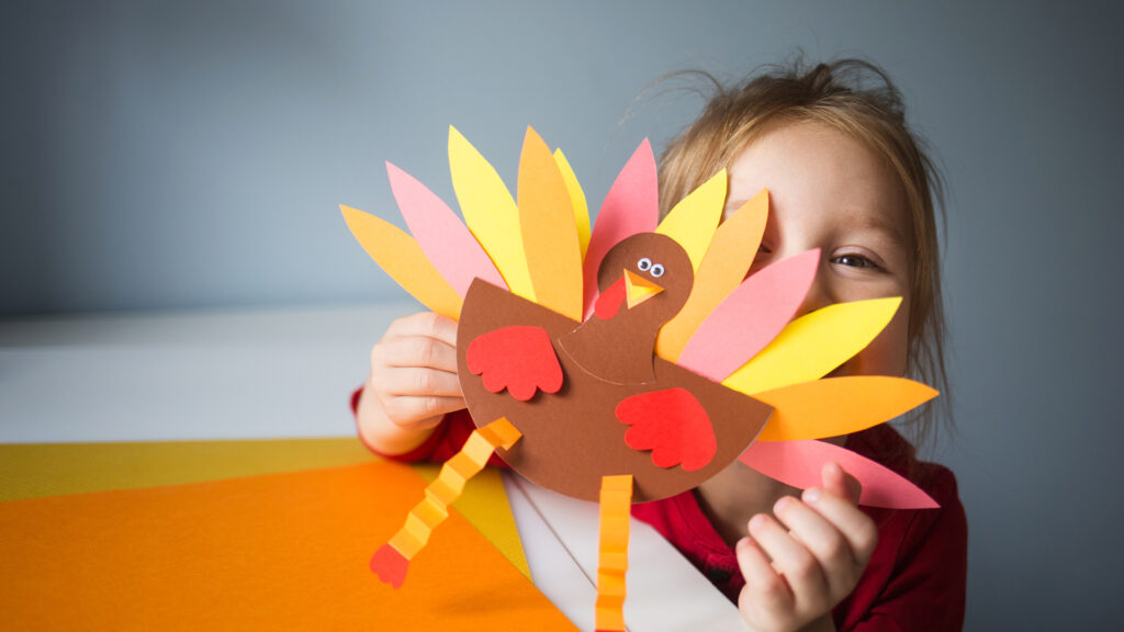 Child holding a handmade paper turkey with colorful feathers, smiling behind it at a table.
