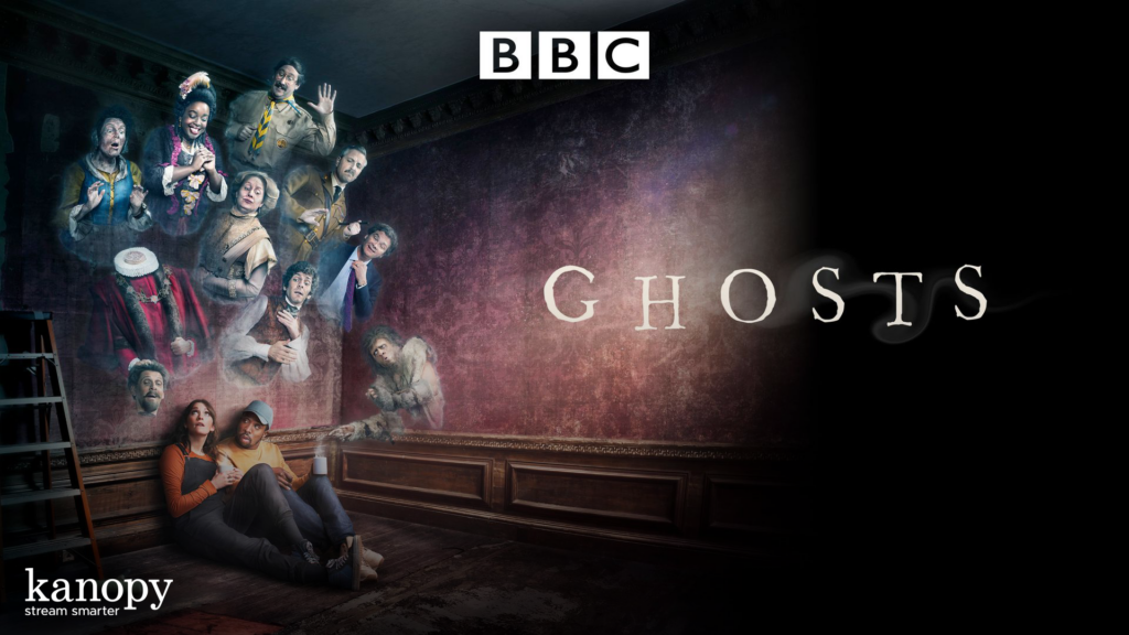 A group of ghosts and two people sit in a dim room with “GHOSTS” and BBC logo above them.