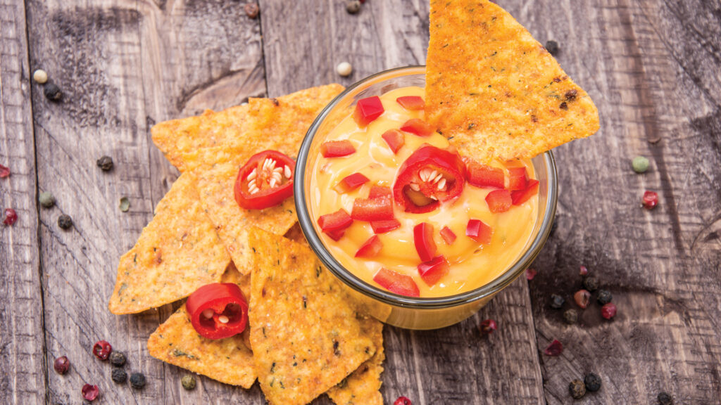 A bowl of cheese dip with red chili slices and tortilla chips on a wooden surface, scattered with peppercorns.