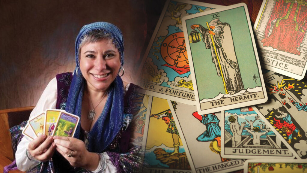 Smiling woman holding tarot cards, with more tarot cards spread out on a table beside her.