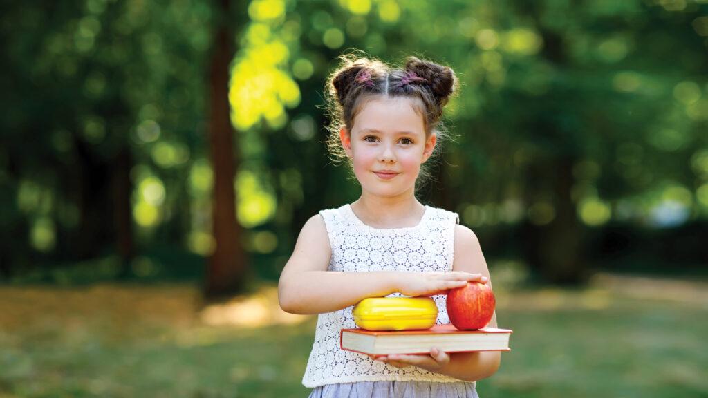 Smiling girl holding books, an apple, and a yellow bell pepper outdoors in a green park.