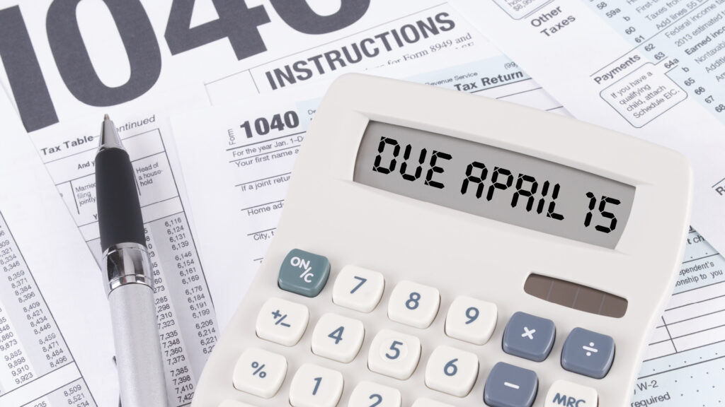 Calculator displays "DUE APRIL 15" on top of IRS 1040 tax forms and a pen.