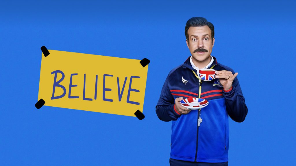 Man in a blue jacket with a cup of tea, next to a yellow poster with "BELIEVE" written on it, against a blue background.