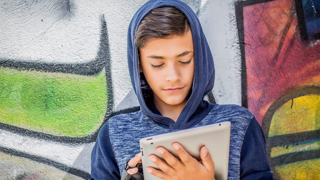 Teen boy in a blue hoodie looking at a tablet, standing in front of a colorful graffiti wall.
