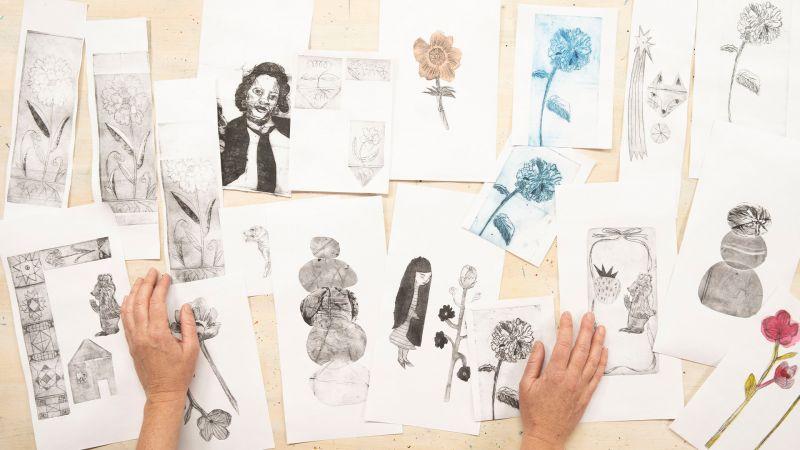 Hands sorting through a collection of black and white and colored flower and portrait sketches on a table.