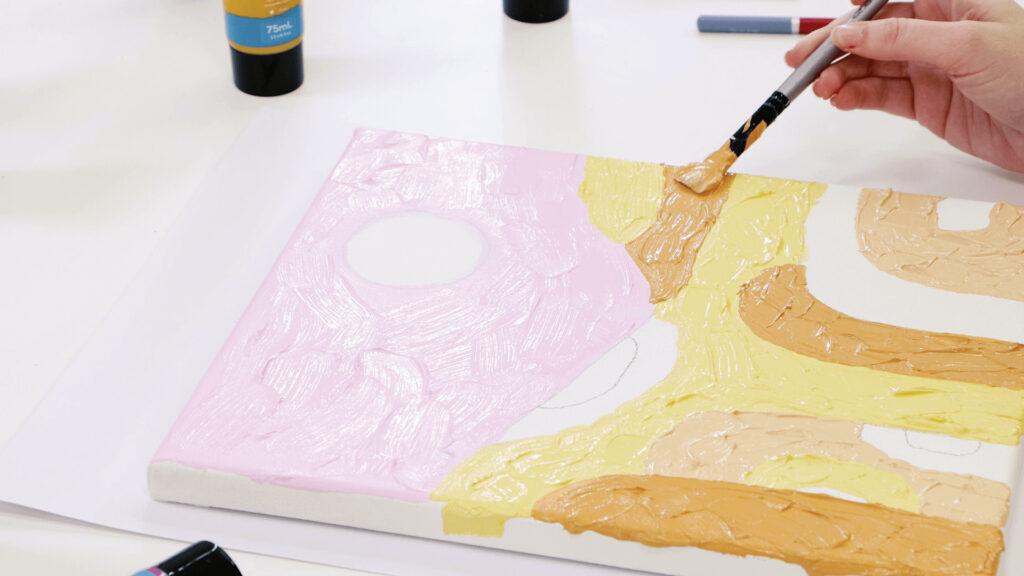 A hand paints abstract shapes with yellow and brown on a canvas next to paint bottles and markers.