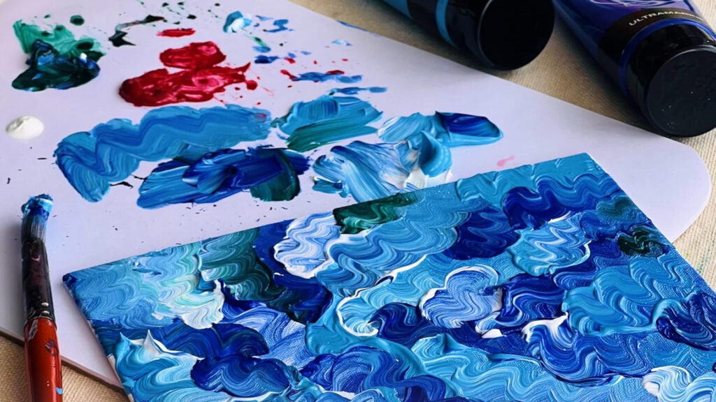 A palette and canvas with blue, white, and red thick swirling paint strokes, next to paint tubes and a brush.