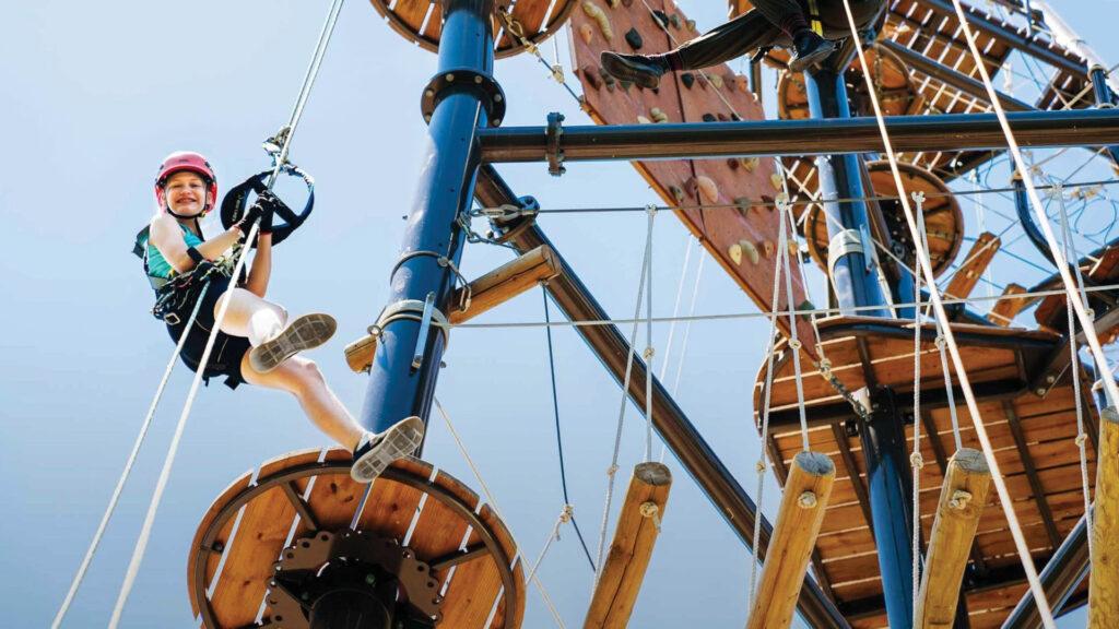 Child in helmet and harness climbing a high ropes course, smiling and holding onto ropes against a clear blue sky.