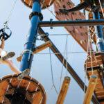 Child in helmet and harness climbing a high ropes course, smiling and holding onto ropes against a clear blue sky.