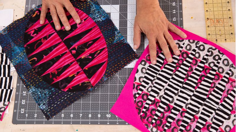 Hands working on colorful fabric quilt designs on a cutting mat, featuring bold pink, black, and white patterns.