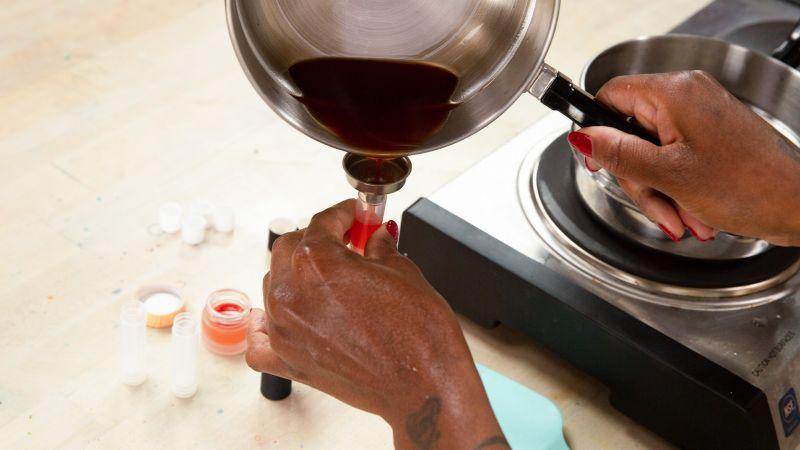 Person pouring liquid into a funnel over a lipstick mold on a counter with small containers nearby.