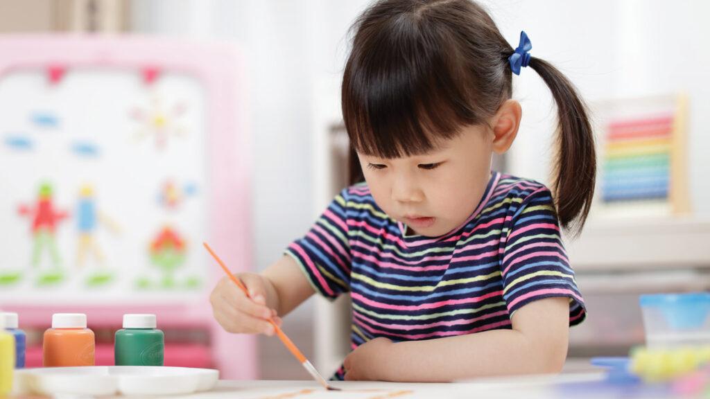 Child painting with a brush at a table, wearing a striped shirt, with art supplies and a colorful easel in the background.