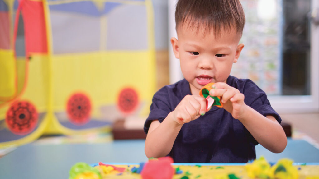 Young child in a navy shirt playing with colorful modeling clay at a table indoors.