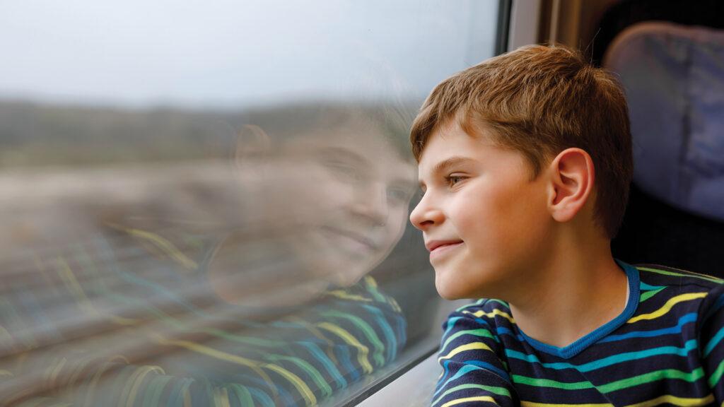 Smiling boy in a striped shirt looking out a train window, with his reflection visible in the glass.
